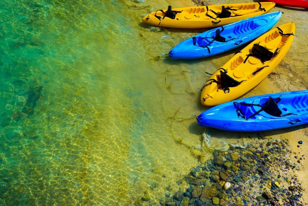 Kayaks in blue and yellow float on clear water by a rocky shore.