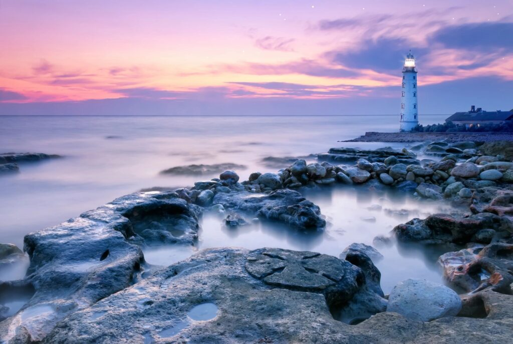 Lighthouse by rocky shore at sunset.